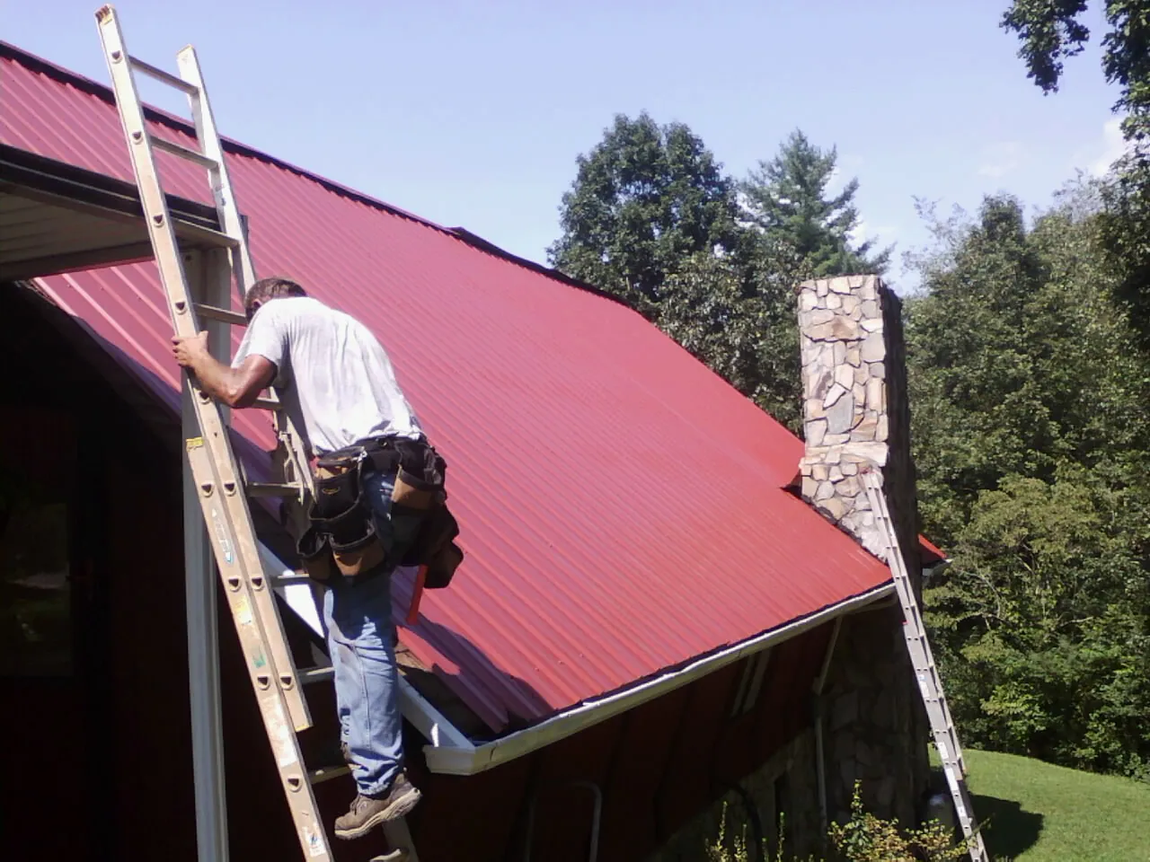 Roofer on a ladder beside a red metal roof and stone chimney — Soffit & Fascia Repair by Roofing Brothers in Palos Verdes Peninsula